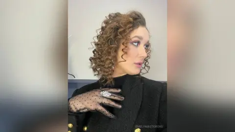 EmelyMastorovo poses in a black blazer and sheer black lace gloves with her curly brown hair framing her face.