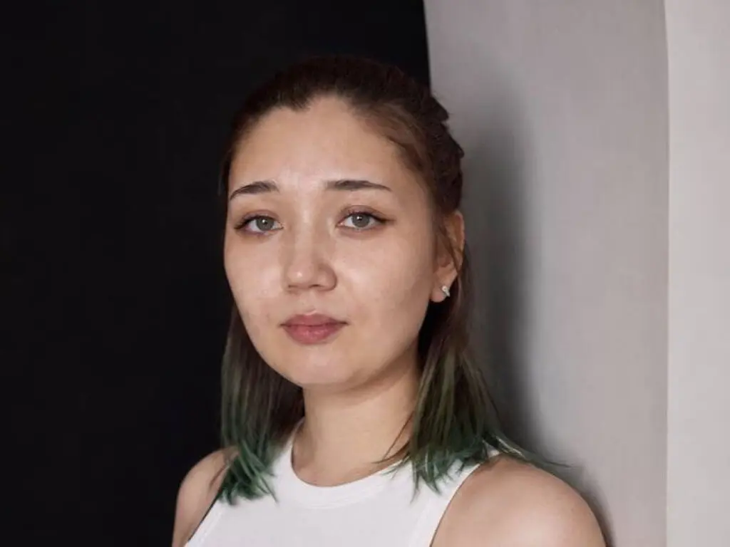 Portrait of a young woman with long dark hair wearing a white tank top against a minimalist black and grey background.