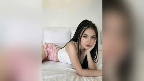 Young woman with long braids in a white tank top relaxing on a white bed in a bright, cozy bedroom.