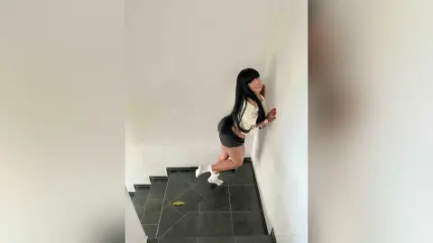 Smiling woman with long black hair in a white top and black shorts posing against a white wall.