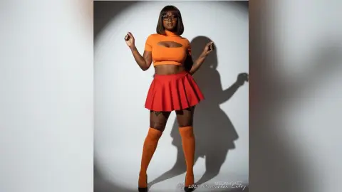 Stylish woman in orange crop top and red mini skirt posing confidently in a studio setting.