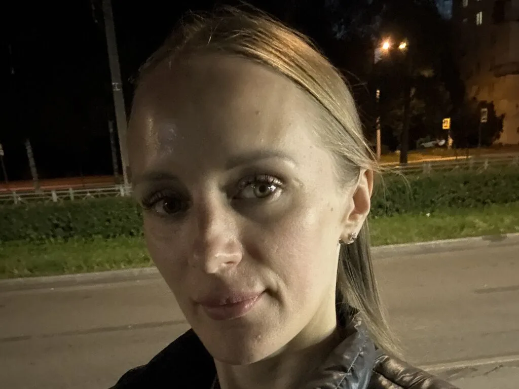MonaLika looks into the camera at night on a street with city lights in the background.