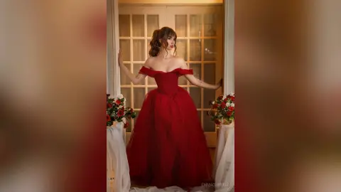 Elegant woman in a flowing red off-the-shoulder ball gown posing by a window in a romantic indoor setting.