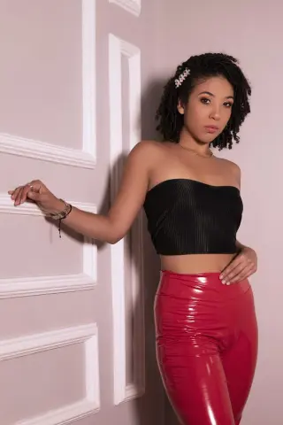 Young woman in a black tube top and red patent leather pants posing against a white paneled wall.