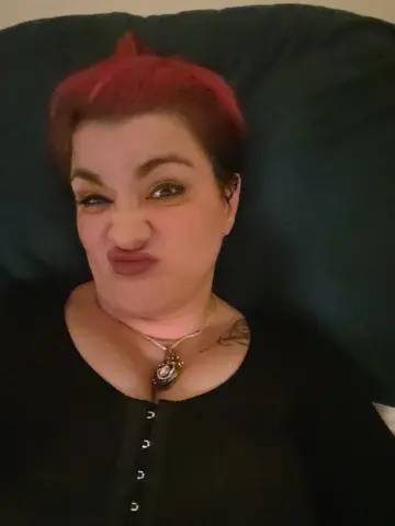 RicanShy pouts her lips and winks, showing her short red hair and silver pendant necklace while wearing a black top.