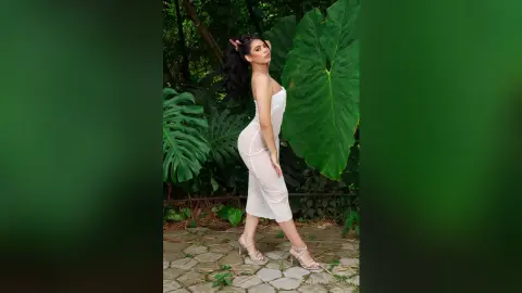 Elegant woman in a white lace midi dress posing amidst lush tropical green leaves in a serene garden setting.