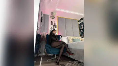 NicoleBella reclines on a blue velvet chair wearing a black bodysuit and sheer black tights.