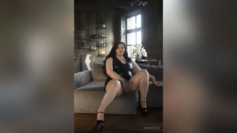 Elegant woman in a black cocktail dress sitting on a grey sofa in a moody, modern living room.