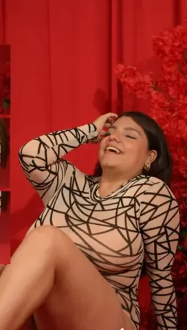 Laughing woman in a black and beige geometric print bodysuit posing against a red curtain background.