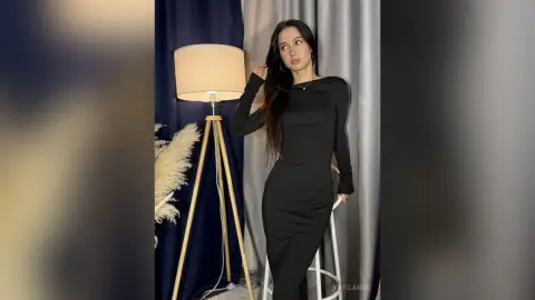 Elegant woman in a sleek black long-sleeve dress posing in a moody, dimly lit indoor setting.
