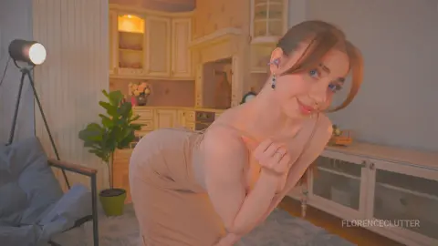 Smiling young woman in a beige dress posing in a cozy, warm-lit modern living room.