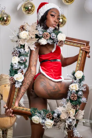 Glamorous woman in red velvet Santa outfit posing with a festive floral frame in a holiday setting.