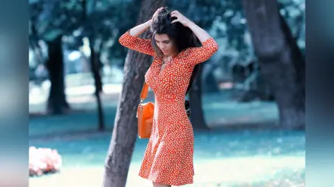 Woman in a red polka dot summer dress carrying an orange bag in a sunny park setting.