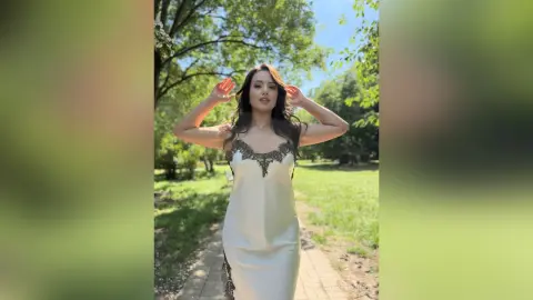 CassyTorres poses outdoors in a park, wearing a white satin slip dress with black lace trim, running her hands through her long dark hair.