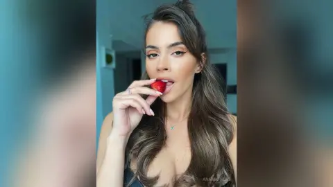 AnaMathers licks a fresh red strawberry while looking into the camera with long wavy hair and a black top.
