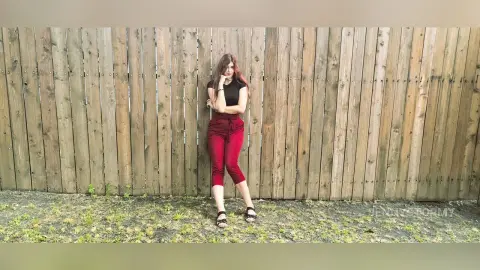 Young woman in black t-shirt and burgundy trousers leaning against a rustic wooden fence in a relaxed mood.