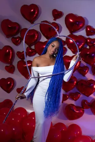 Smiling woman in a white off-the-shoulder dress holding a heart prop amidst red heart balloons for Valentine's Day.