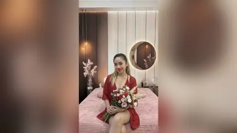 Smiling woman in a red dress holding a bouquet of flowers in a modern, cozy bedroom setting.