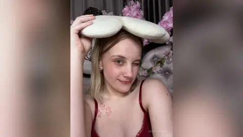 Smiling blonde woman in a red camisole holding a white pillow on her head in a cozy bedroom setting.