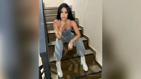 Stylish woman in denim jumpsuit and white boots sitting on indoor stairs with a confident mood.
