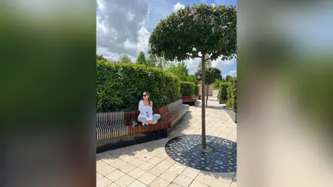 Woman in white summer outfit sitting on a park bench under a sunny sky in a lush green garden.