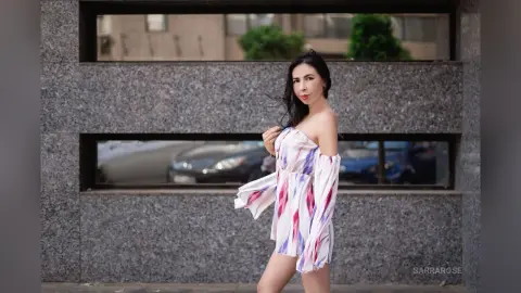 Elegant woman in a white floral off-the-shoulder dress posing outdoors against a stone building background.