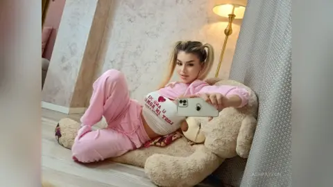 AshPayton wearing a pink crop top and pink sweatpants, lying on a large teddy bear in a room with a lamp and grey curtain.