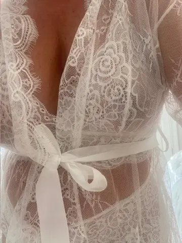 Woman in a delicate white lace sheer robe with a satin waist tie in a bright, airy setting.