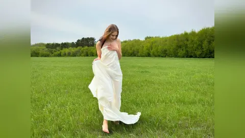 Dreamy woman in a flowing white dress walking through a lush green meadow under a soft sky.