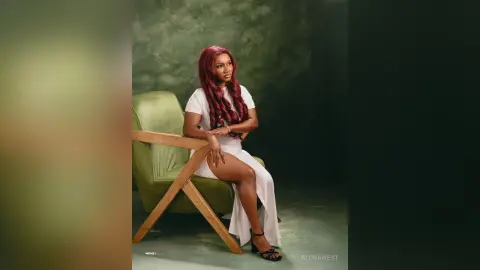 Elegant woman with red hair in a white dress sitting on a green velvet chair against a dark studio background.