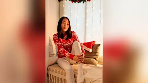 Smiling woman in a red festive sweater sitting on a bed near holiday lights in a cozy winter setting.