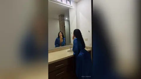 Young woman in a blue satin robe applying makeup in a modern bathroom mirror.
