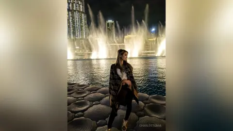 Stylish woman in a patterned cardigan posing by the Dubai Fountain at night with city lights reflecting on water.