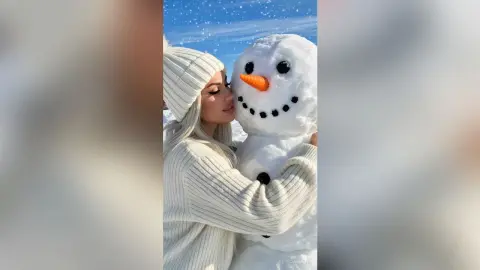 Smiling woman in a white knit sweater hugging a happy snowman in a sunny winter snow landscape.