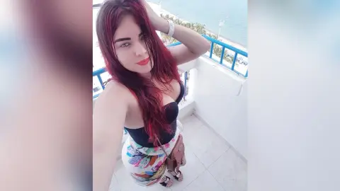 Young woman with red hair in a black tank top posing on a sunny white balcony overlooking the ocean.