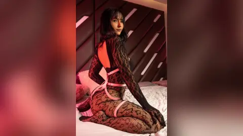 Young woman in black lace bodysuit sitting on a bed in a moody, dimly lit bedroom setting.