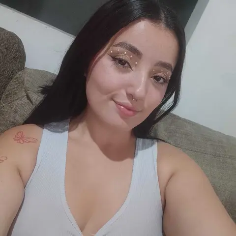 Smiling young woman with facial freckle makeup wearing a white ribbed tank top in a cozy indoor setting.