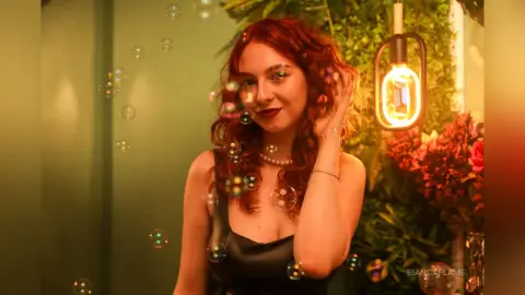 Redheaded woman in a black dress surrounded by floating bubbles in a warm, dreamy indoor setting.