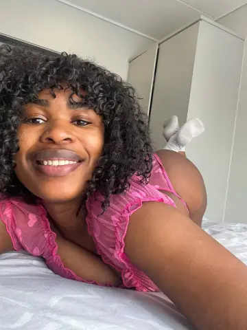 Smiling woman with curly hair wearing pink lace lingerie while relaxing on a white bed in a bright bedroom.