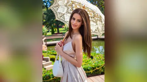 Elegant woman in a white sundress holding a lace parasol in a sunny garden setting.