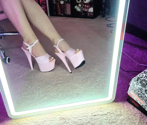 Close-up of pink high heel platform sandals worn by a person standing on a carpeted floor near a lighted mirror.