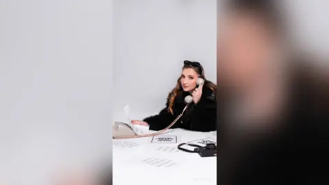 Smiling woman in a black faux fur coat talking on a vintage telephone at a white desk in a bright studio.