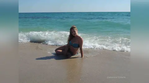 Young woman in a teal bikini relaxing on a sunny beach with ocean waves crashing in the background.