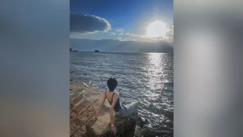 Young woman in a black tank top sitting on rocks by the ocean under a bright sunset sky.