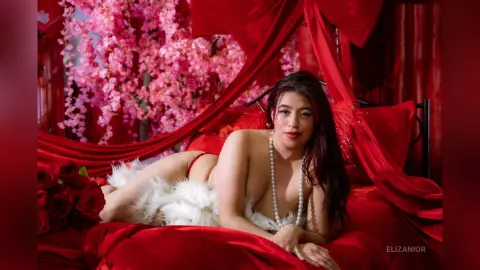 Beautiful woman in white feather lingerie and pearls lounging on a red bed amidst pink cherry blossoms.