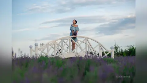 Elegant woman in a blue dress holding flowers on a white bridge amidst a dreamy lavender field at sunset.