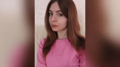 Young woman with long brown hair wearing a pink sweatshirt in a bright indoor setting with a calm expression.