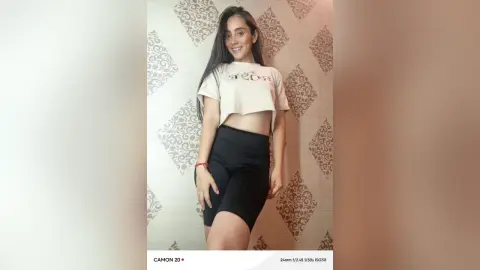 Smiling young woman in a white crop top and black biker shorts posing against a patterned wallpaper background.