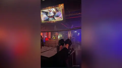 Person relaxing in a dark nightclub with feet up on a table near a glowing television screen.