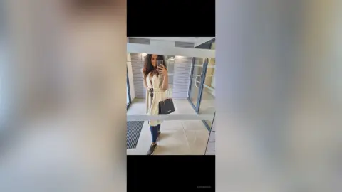 Stylish woman in a beige trench coat and jeans taking a mirror selfie in a modern bright hallway.
